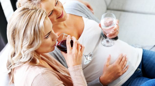 Eine schwangere Frau mit Wasserglas und eine nicht schwangere Frau mit Weinglas sitzen nebeneinander auf einem Sofa.