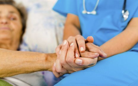 Man sieht eine ältere Frau im Krankenbett und eine Krankenschwester, die am Bett sitzt.