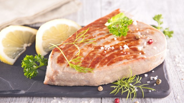 Auf einem Holztisch liegt eine Servierplatte mit einem Thunfischfilet, garniert mit Zitronenstücken, Kräutern udn Gewürzen.