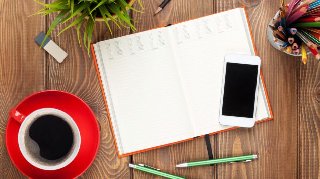 Ein offener Kalender, daneben ein Smartphone, Stifte und eine Tasse Kaffee